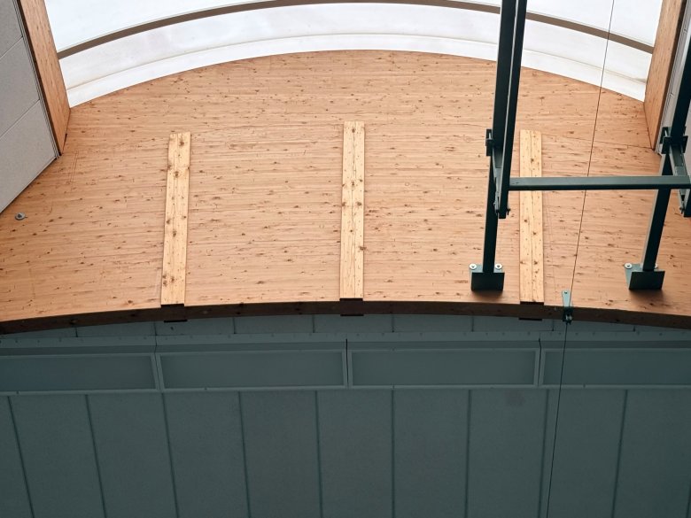 Blick nach oben auf die Holzkonstruktion und das geschwungene Dach einer Sporthalle. Die Dachfläche besteht aus hellen Holzelementen und ist teilweise von lichtdurchlässigen Oberlichtern überspannt. Im Vordergrund sind Teile einer schwarzen technischen Konstruktion und eine Aufhängung zu sehen.