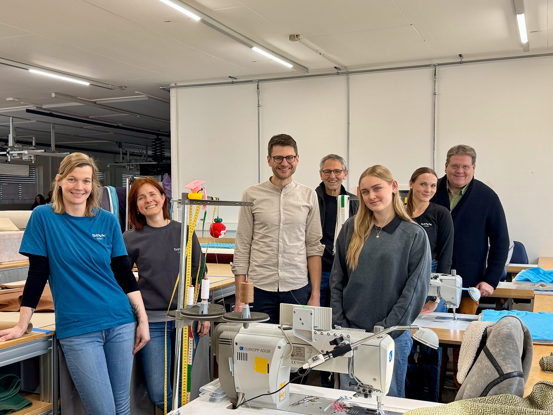 Sieben Personen stehen in einer hellen Polsterei-Werkstatt zwischen Arbeitstischen und Nähmaschinen und lächeln in die Kamera. Im Vordergrund befindet sich eine große Industrienähmaschine mit Garnrollen; daneben liegen Stoffe und Werkzeuge. Hinter der Gruppe sind weitere Näharbeitsplätze, Maßbänder und Polstermaterialien zu sehen. Einige tragen Arbeitskleidung mit Firmenlogo. Die Umgebung wirkt ordentlich und professionell.