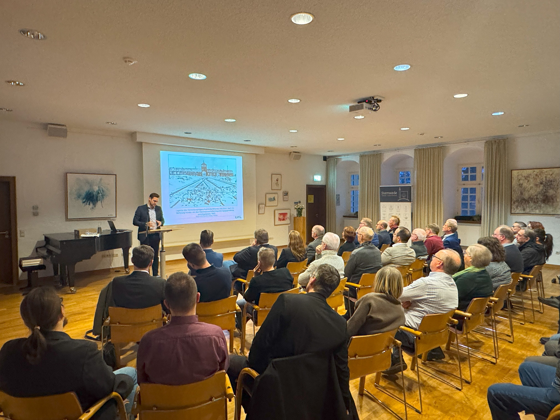In einem Veranstaltungsraum hält ein Mann an einem Rednerpult einen Vortrag vor sitzendem Publikum. Die Zuhörerinnen und Zuhörer sitzen in mehreren Reihen und blicken auf eine projizierte Präsentationsfolie. Auf der Folie ist eine historische Darstellung zu sehen. Der Raum ist hell beleuchtet, an den Wänden hängen Bilder.