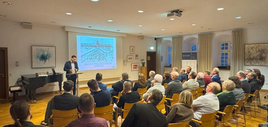 In einem Veranstaltungsraum hält ein Mann an einem Rednerpult einen Vortrag vor sitzendem Publikum. Die Zuhörerinnen und Zuhörer sitzen in mehreren Reihen und blicken auf eine projizierte Präsentationsfolie. Auf der Folie ist eine historische Darstellung zu sehen. Der Raum ist hell beleuchtet, an den Wänden hängen Bilder.