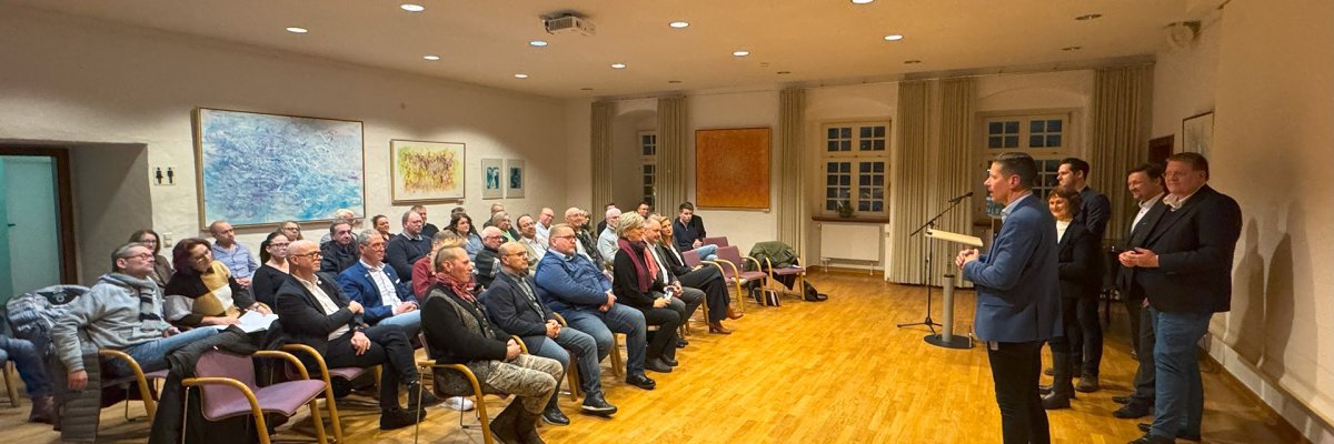 In einem Veranstaltungsraum stehen fünf Personen vor sitzendem Publikum. Die Zuhörerinnen und Zuhörer sitzen in mehreren Reihen und blicken auf eine projizierte Präsentationsfolie. Der Raum ist hell beleuchtet, an den Wänden hängen Bilder.