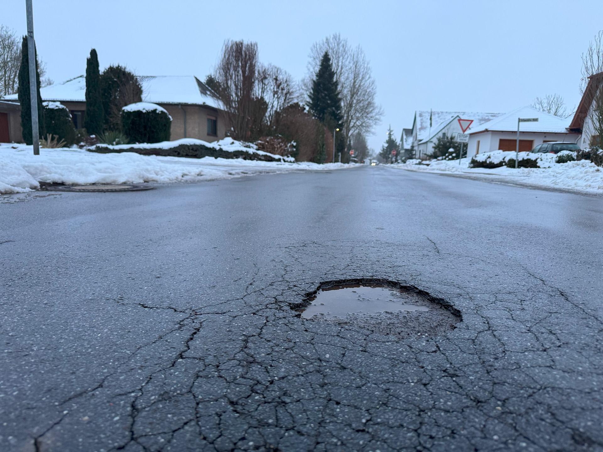 Nasse Wohnstraße im Winter mit Schnee am Straßenrand. Im Vordergrund ein rundes Schlagloch mit Wasser, umgeben von Rissen im Asphalt. Im Hintergrund Einfamilienhäuser, Bäume ohne Blätter und ein Stoppschild an einer Kreuzung.