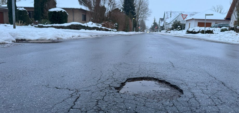 Nasse Wohnstraße im Winter mit Schnee am Straßenrand. Im Vordergrund ein rundes Schlagloch mit Wasser, umgeben von Rissen im Asphalt. Im Hintergrund Einfamilienhäuser, Bäume ohne Blätter und ein Stoppschild an einer Kreuzung.