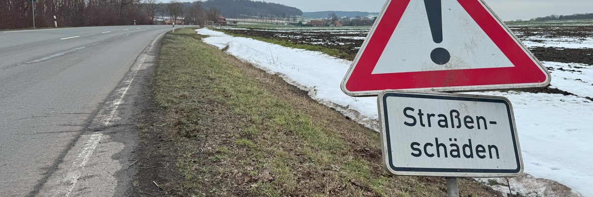 Landstraße im Winter mit Schnee auf den Feldern. Am rechten Straßenrand steht ein dreieckiges Warnschild mit Ausrufezeichen und dem Zusatz „Straßenschäden“. Die Straße verläuft leicht gebogen durch eine ländliche Landschaft mit Bäumen und Hügeln im Hintergrund.
