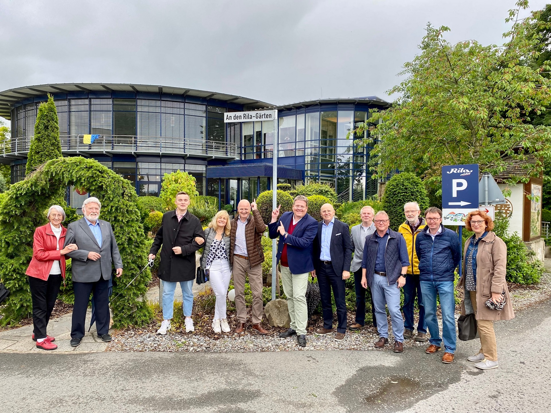Eine Gruppe von zwölf Erwachsenen steht nebeneinander vor einem modernen, zweigeschossigen Gebäude mit viel Glas und runder Architektur. Über dem Eingang hängt ein Schild mit der Aufschrift „An den Rila-Gärten“. Die Personen, überwiegend mittleren bis höheren Alters, lächeln in die Kamera; einige stehen eng beieinander oder haben die Arme eingehakt. Ein Mann in der Mitte zeigt mit beiden Händen auf das Straßenschild. Rechts im Bild steht ein blaues Parkplatzschild mit einem „P“ und dem Zusatz „Rila“. Im Vordergrund sind Asphalt und eine kleine Pfütze zu sehen, im Hintergrund Bäume, Sträucher und ein bewölkter Himmel.