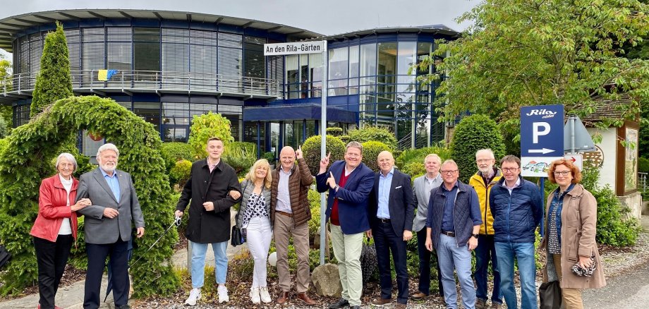 Enthüllung "In den Rila-Gärten" Eine Gruppe von zwölf Erwachsenen steht nebeneinander vor einem modernen, zweigeschossigen Gebäude mit viel Glas und runder Architektur. Über dem Eingang hängt ein Schild mit der Aufschrift „An den Rila-Gärten“. Die Personen, überwiegend mittleren bis höheren Alters, lächeln in die Kamera; einige stehen eng beieinander oder haben die Arme eingehakt. Ein Mann in der Mitte zeigt mit beiden Händen auf das Straßenschild. Rechts im Bild steht ein blaues Parkplatzschild mit einem „P“ und dem Zusatz „Rila“. Im Vordergrund sind Asphalt und eine kleine Pfütze zu sehen, im Hintergrund Bäume, Sträucher und ein bewölkter Himmel.