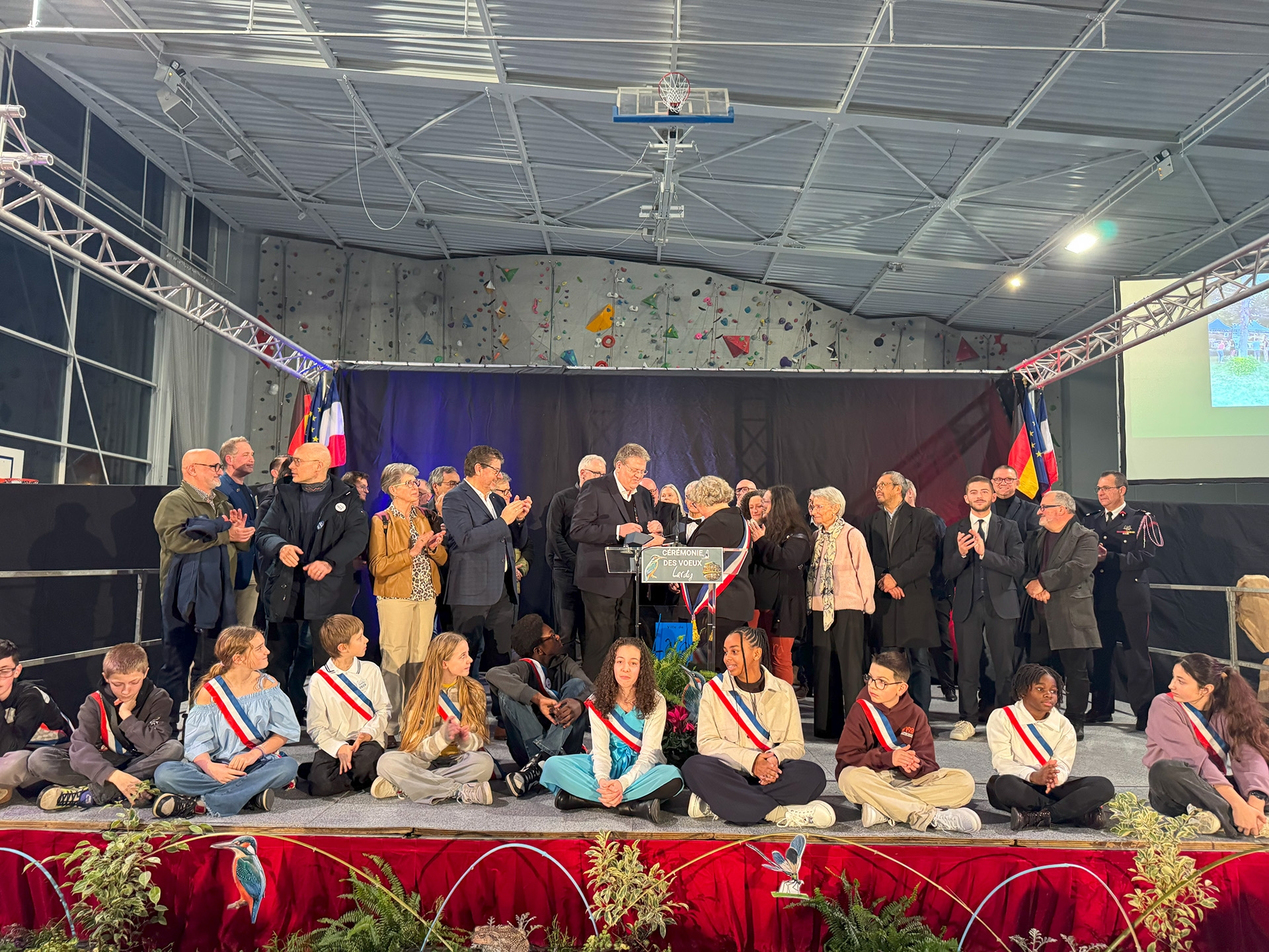 Auf einer großen Bühne in einer Sporthalle stehen und sitzen zahlreiche Menschen bei einer feierlichen Veranstaltung. Im Vordergrund sitzen Kinder und Jugendliche im Schneidersitz auf der Bühne, viele tragen Schärpen in den Farben Blau, Weiß und Rot. Hinter ihnen stehen Erwachsene, überwiegend in dunkler Kleidung, einige applaudieren. In der Mitte befindet sich ein Rednerpult mit der Aufschrift „Cérémonie des Vœux“, davor Blumen. Im Hintergrund hängen französische und deutsche Flaggen sowie eine dunkle Bühnenkulisse. Über der Bühne ist die Hallendecke mit einem Basketballkorb und einer Kletterwand zu sehen. Die Atmosphäre wirkt festlich und würdevoll.