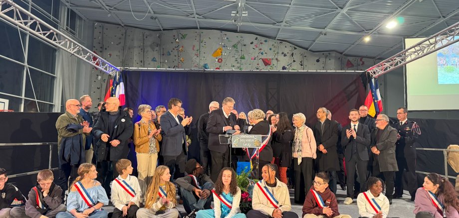Lardy 2026 Auf einer großen Bühne in einer Sporthalle stehen und sitzen zahlreiche Menschen bei einer feierlichen Veranstaltung. Im Vordergrund sitzen Kinder und Jugendliche im Schneidersitz auf der Bühne, viele tragen Schärpen in den Farben Blau, Weiß und Rot. Hinter ihnen stehen Erwachsene, überwiegend in dunkler Kleidung, einige applaudieren. In der Mitte befindet sich ein Rednerpult mit der Aufschrift „Cérémonie des Vœux“, davor Blumen. Im Hintergrund hängen französische und deutsche Flaggen sowie eine dunkle Bühnenkulisse. Über der Bühne ist die Hallendecke mit einem Basketballkorb und einer Kletterwand zu sehen. Die Atmosphäre wirkt festlich und würdevoll.