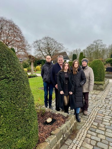 Fünf Personen stehen dicht beieinander auf einem gepflasterten Weg auf einem Friedhof. Sie blicken ruhig und ernst in die Kamera und tragen winterliche Kleidung wie Mäntel und Jacken. Im Vordergrund links befindet sich ein kleines, rechteckiges Beet mit dunkler Erde; darin liegt ein ovaler, beschrifteter Gedenkstein auf einer schwarzen Platte. Umgeben ist das Beet von niedrigen Steinmauern und sorgfältig geschnittenen grünen Sträuchern. Im Hintergrund sind weitere Grabstätten, Bäume ohne Blätter und ein Gebäude zu sehen. Der Himmel ist grau und bewölkt, die Atmosphäre wirkt still und würdevoll.