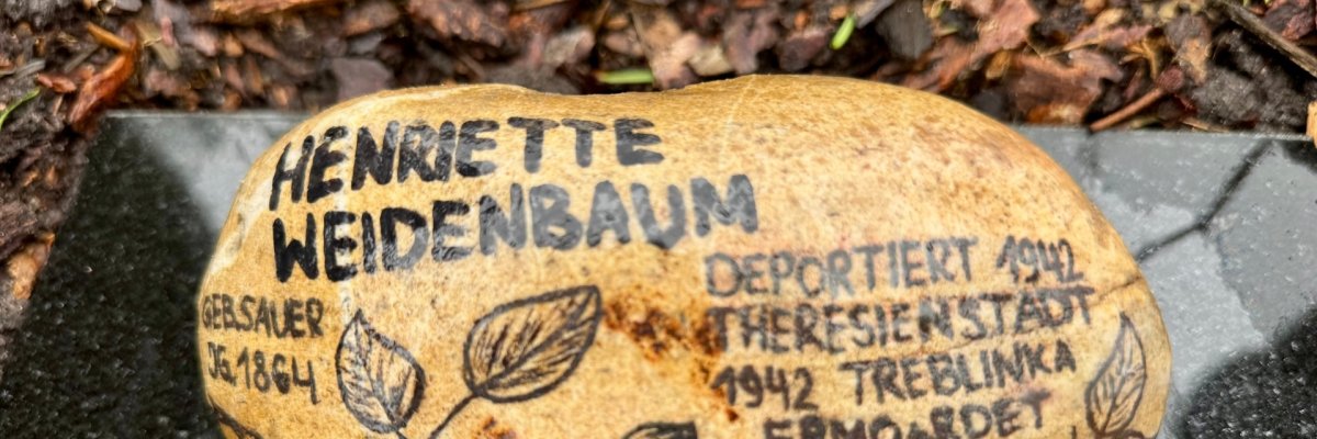 Ein ovaler, hellbrauner Gedenkstein liegt auf einer dunklen Steinplatte im Freien, umgeben von feuchtem Laub und Erde. Auf dem Stein stehen handschriftlich wirkende schwarze Inschriften und florale Zeichnungen. Lesbarer Text: „Henriette Weidenbaum“, links „Gersauer Nr. 18 CH“, rechts „Deportiert 1942 Theresienstadt 1942 Treblinka ermordet“. Der Stein ist mit gezeichneten Blumen und Blättern verziert und wirkt wie ein persönliches Erinnerungszeichen an ein Opfer des Holocaust.