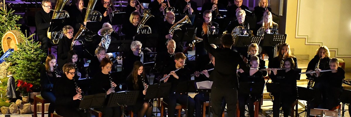 Großes Blasorchester bei einem Konzert in einem festlich beleuchteten Saal. Zahlreiche Musikerinnen und Musiker in schwarzer Kleidung sitzen und spielen Holz- und Blechblasinstrumente. In der Mitte steht ein Dirigent mit erhobenen Armen und leitet das Orchester. Im Hintergrund sind Kerzenleuchter und farbiges Bühnenlicht zu sehen, links ein geschmückter Weihnachtsbaum. Im Vordergrund erkennt man die Silhouetten des Publikums.