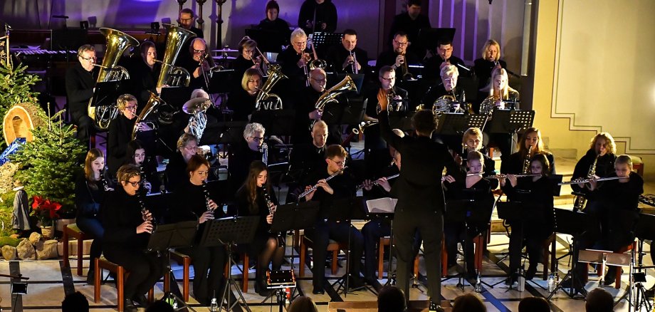 Blasorchester Handorf-Langenberg Großes Blasorchester bei einem Konzert in einem festlich beleuchteten Saal. Zahlreiche Musikerinnen und Musiker in schwarzer Kleidung sitzen und spielen Holz- und Blechblasinstrumente. In der Mitte steht ein Dirigent mit erhobenen Armen und leitet das Orchester. Im Hintergrund sind Kerzenleuchter und farbiges Bühnenlicht zu sehen, links ein geschmückter Weihnachtsbaum. Im Vordergrund erkennt man die Silhouetten des Publikums.