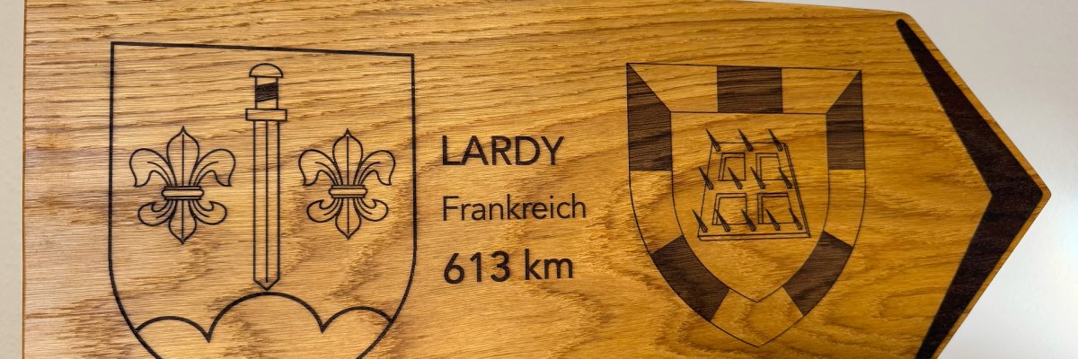 Holschild Kurpark Nahaufnahme eines hölzernen Wegweiserschildes aus massivem Holz. Das Schild zeigt nach rechts und ist auf einem senkrechten Holzpfosten befestigt. In das Holz sind zwei Wappen eingraviert: links ein Wappen mit einem Schwert und zwei Lilien, rechts ein Wappenschild mit einem stilisierten Gebäude. In der Mitte steht der Text „LARDY – Frankreich – 613 km“. Das Schild wirkt handgefertigt und sorgfältig bearbeitet.