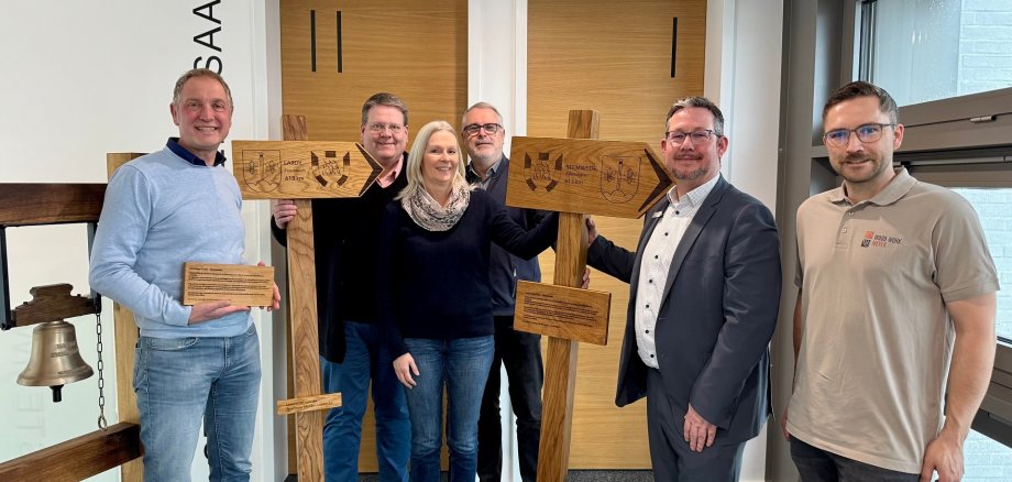 Sparkassensponsoring Partnerschaftsverein Sechs erwachsene Personen stehen nebeneinander in einem hellen Innenraum. In der Mitte halten zwei Personen jeweils ein großes, hölzernes Wegweiserschild in Kreuzform. Die Schilder sind handgefertigt und mit eingravierten Wappen, Ortsnamen und Entfernungsangaben versehen. Links im Bild steht zusätzlich eine hölzerne Konstruktion mit einer Glocke. Alle Personen lächeln in die Kamera und tragen überwiegend legere bis halbformelle Kleidung. Der Raum wirkt modern, mit hellen Wänden, Holzflächen und großen Fenstern auf der rechten Seite.