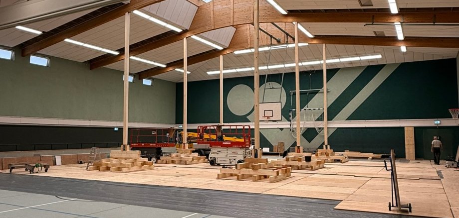 Dachsanierung Dielingen Blick in eine Sporthalle während der Sanierung der Dachkonstruktion. Mehrere Dachträger werden mit senkrechten Holzstützen gesichert. Auf dem Hallenboden liegen Holzbauteile, Arbeitsmaterialien und zwei Hebebühnen. Teile der Deckenverkleidung wurden geöffnet, um die Dachträger instand zu setzen. Im Hintergrund sind Basketballkörbe und die Hallenwände zu sehen.