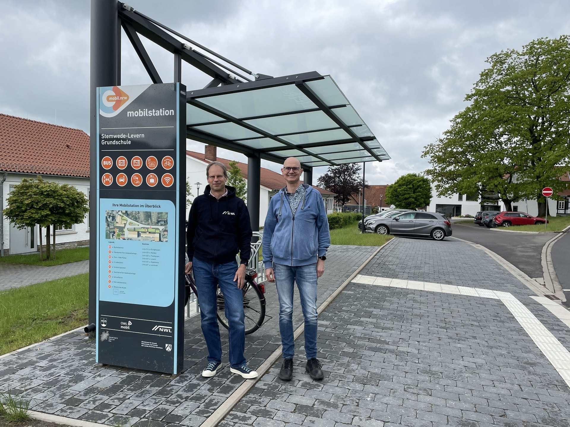 Zwei Personen stehen unter einem überdachten Mobilitätsknoten neben einer Informationsstele mit der Aufschrift „mobilstation“. Auf der Stele sind Symbole für verschiedene Mobilitätsangebote wie Bus, Fahrrad und Parken sowie ein Übersichtsplan zu sehen. Hinter ihnen befindet sich ein Fahrradständer. Die Mobilstation liegt an einer gepflasterten Straße mit einem taktilen Leitsystem für blinde und sehbehinderte Menschen. Im Hintergrund sind Bäume, geparkte Autos und Gebäude eines Dorfes unter einem bewölkten Himmel zu sehen.