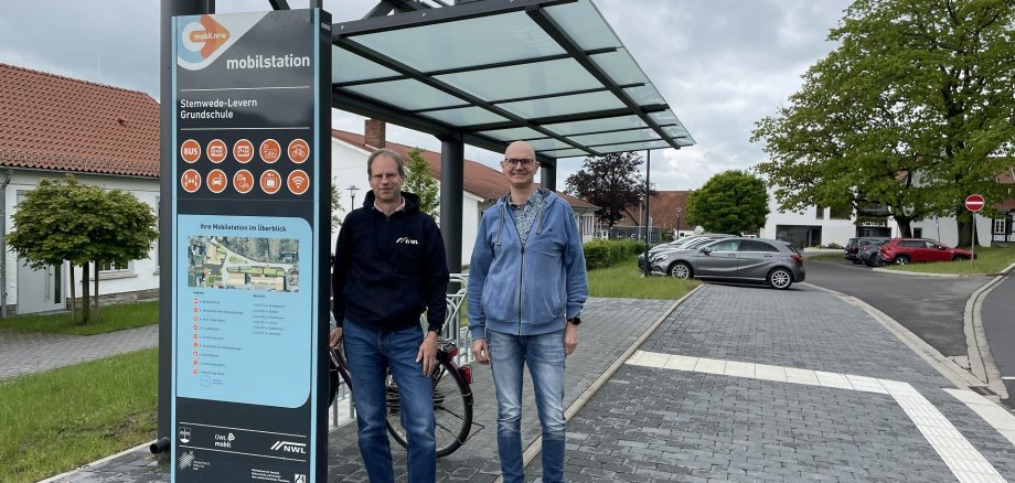 Zwei Personen stehen unter einem überdachten Mobilitätsknoten neben einer Informationsstele mit der Aufschrift „mobilstation“. Auf der Stele sind Symbole für verschiedene Mobilitätsangebote wie Bus, Fahrrad und Parken sowie ein Übersichtsplan zu sehen. Hinter ihnen befindet sich ein Fahrradständer. Die Mobilstation liegt an einer gepflasterten Straße mit einem taktilen Leitsystem für blinde und sehbehinderte Menschen. Im Hintergrund sind Bäume, geparkte Autos und Gebäude eines Dorfes unter einem bewölkten Himmel zu sehen.
