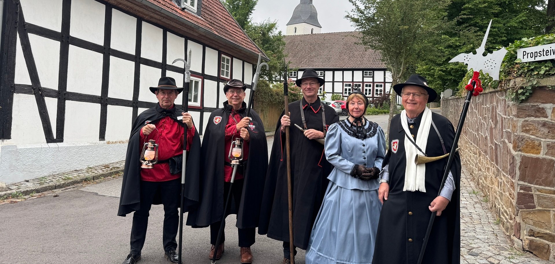 Fünf Personen in historischer Nachtwächterkleidung stehen auf einer ruhigen Dorfstraße vor Fachwerkhäusern. Vier tragen schwarze Umhänge, Hüte und traditionelle Nachtwächterausrüstung wie Hellebarden, Laternen und ein Signalhorn. Eine Person trägt ein hellblaues historisches Kleid mit schwarzem Schal und Handschuhen. Im Hintergrund sind weitere Fachwerkgebäude und der Kirchturm zu sehen, eingerahmt von großen Bäumen. Rechts weist ein Schild auf die Propstei hin. Die Aufnahme vermittelt eine historische Atmosphäre.