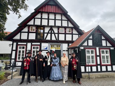 Sechs Personen stehen vor einem historischen Fachwerkgebäude mit weißer Fassade und schwarzen Holzbalken. Fünf von ihnen tragen traditionelle Nachtwächterkleidung mit schwarzen Umhängen, Hüten sowie Ausrüstung wie Laternen, Hellebarden und einem Signalhorn. Eine Person trägt ein hellblaues historisches Kleid mit schwarzem Schal und Handschuhen. Daneben steht eine Frau in moderner heller Kleidung. Über dem Eingang hängt ein Schild der Gemeinde Stemwede, an der Tür ist eine Nachtwächter-Figur als Dekoration angebracht. Das Gruppenfoto vermittelt den Eindruck eines offiziellen Empfangs vor dem historischen Rathaus.