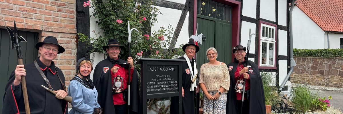 Sechs Personen stehen vor einem historischen Fachwerkgebäude mit gepflegtem Vorgarten. Fünf tragen traditionelle Nachtwächterkleidung mit schwarzen Umhängen und Hüten sowie Ausrüstung wie Hellebarden, Laternen und einem Signalhorn. Eine Person trägt ein hellblaues historisches Kleid mit schwarzem Schal und Handschuhen. Daneben steht eine Frau in moderner heller Kleidung. Im Vordergrund informiert eine Tafel über den „Alten Ausspann“, umgeben von Rosen, Lavendel und weiteren Pflanzen. Das Gruppenfoto vermittelt den Eindruck eines gemeinsamen Besuchs an einem historischen Ort.