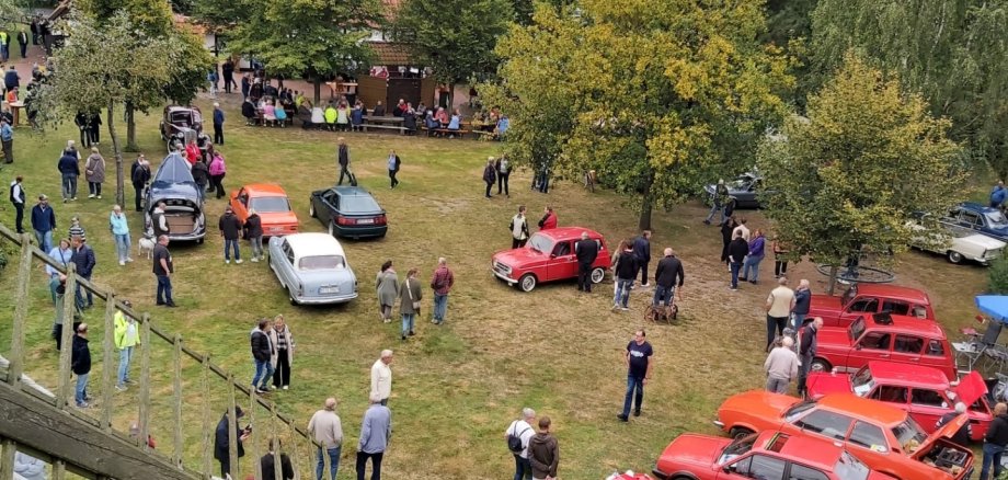 Oldtimertreffen Oppenwehe 2025 Blick von einem erhöhten Standpunkt auf das Gelände eines Oldtimertreffens. Auf einer großen Wiese sind zahlreiche historische Autos in verschiedenen Farben und Bauformen ausgestellt. Viele Besucherinnen und Besucher schlendern zwischen den Fahrzeugen, betrachten die Oldtimer oder kommen miteinander ins Gespräch. Im Hintergrund stehen große Bäume und ein Gebäude mit Sitzgelegenheiten, an denen weitere Gäste verweilen. Links im Vordergrund ist ein Teil der Flügel einer historischen Windmühle zu sehen, wodurch die Aufnahme einen Überblick über das gesamte Veranstaltungsgelände vermittelt.