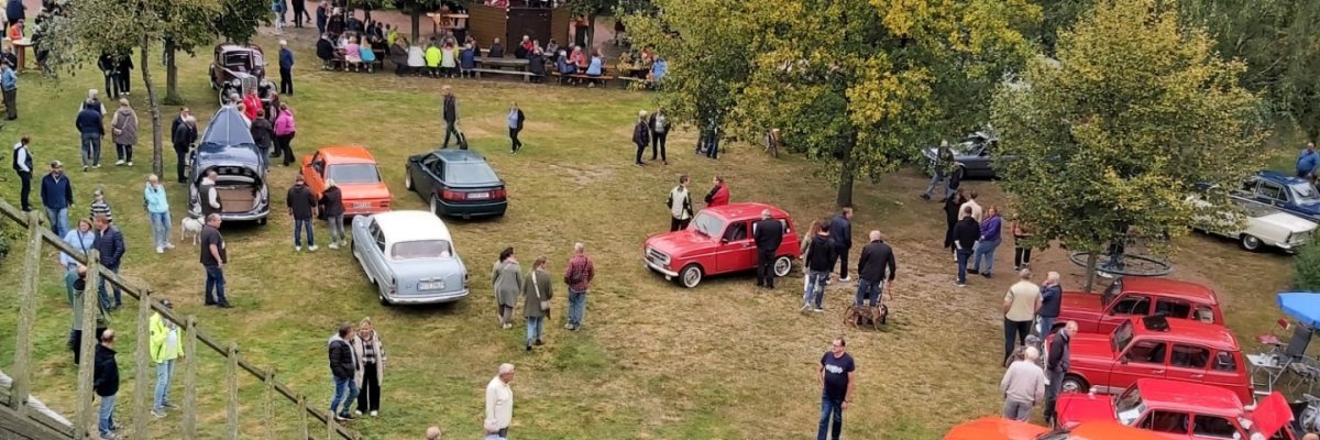 Oldtimertreffen Oppenwehe 2025 Blick von einem erhöhten Standpunkt auf das Gelände eines Oldtimertreffens. Auf einer großen Wiese sind zahlreiche historische Autos in verschiedenen Farben und Bauformen ausgestellt. Viele Besucherinnen und Besucher schlendern zwischen den Fahrzeugen, betrachten die Oldtimer oder kommen miteinander ins Gespräch. Im Hintergrund stehen große Bäume und ein Gebäude mit Sitzgelegenheiten, an denen weitere Gäste verweilen. Links im Vordergrund ist ein Teil der Flügel einer historischen Windmühle zu sehen, wodurch die Aufnahme einen Überblick über das gesamte Veranstaltungsgelände vermittelt.