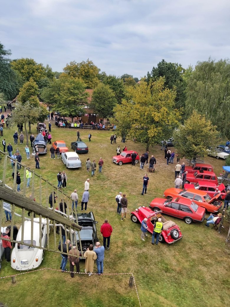 Oldtimertreffen Oppenwehe 2025 Blick von einem erhöhten Standpunkt auf das Gelände eines Oldtimertreffens. Auf einer großen Wiese sind zahlreiche historische Autos in verschiedenen Farben und Bauformen ausgestellt. Viele Besucherinnen und Besucher schlendern zwischen den Fahrzeugen, betrachten die Oldtimer oder kommen miteinander ins Gespräch. Im Hintergrund stehen große Bäume und ein Gebäude mit Sitzgelegenheiten, an denen weitere Gäste verweilen. Links im Vordergrund ist ein Teil der Flügel einer historischen Windmühle zu sehen, wodurch die Aufnahme einen Überblick über das gesamte Veranstaltungsgelände vermittelt.