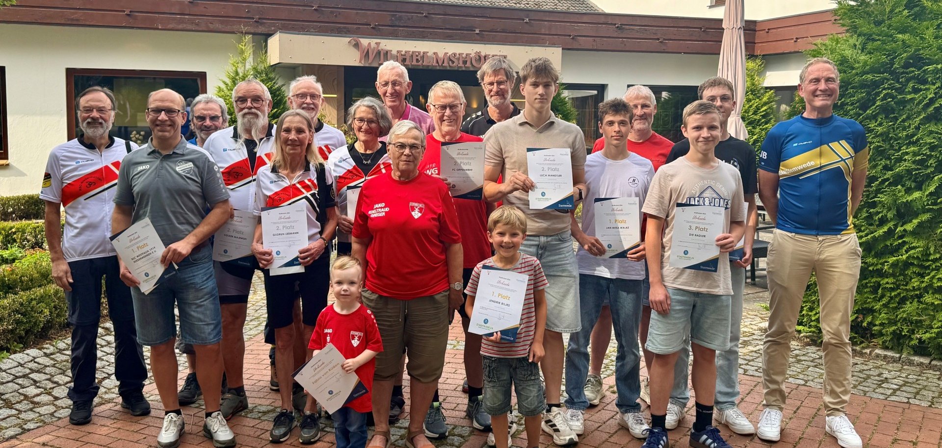 Gruppenfoto vor der Wilhelmshöhe: Rund 20 Teilnehmerinnen und Teilnehmer des STADTRADELNs stehen mit Urkunden für die Ehrung der Gemeinde Stemwede zusammen. Vertreten sind Kinder, Jugendliche und Erwachsene, viele tragen Fahrradtrikots oder Sportkleidung. Vorne stehen zwei Kinder mit Urkunden, dahinter die ausgezeichneten Radlerinnen und Radler in mehreren Reihen. Im Hintergrund sind das Gebäude der Wilhelmshöhe sowie ein bewölkter Himmel zu sehen. Die Stimmung wirkt stolz und fröhlich.