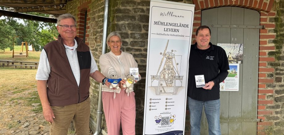 Drei Personen stehen vor der historischen Holthoffschen Hofmahlmühle auf dem Mühlengelände in Levern. Zwischen ihnen befindet sich ein Roll-up mit einer Zeichnung der Mühle und Informationen zum neuen digitalen Mühlenchatbot. Die Person in der Mitte hält die Maskottchen Stemmi und Wedi sowie eine Infokarte, die beiden Personen links und rechts präsentieren ebenfalls Informationskarten zum Angebot. Im Hintergrund sind die Natursteinfassade der Mühle, eine Holztür mit Infotafel sowie Teile des Außengeländes mit Bäumen und Sitzgelegenheiten zu sehen.