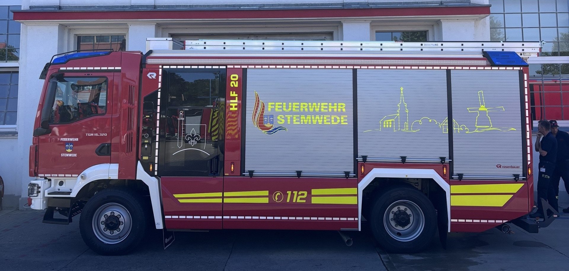 Ein rotes Hilfeleistungslöschgruppenfahrzeug (HLF 20) der Feuerwehr Stemwede steht vor dem Werk des Feuerwehrfahrzeugherstellers Rosenbauer. Auf der Fahrzeugseite sind das Wappen der Gemeinde Stemwede, die Aufschrift „Feuerwehr Stemwede“ sowie eine stilisierte Silhouette mit Kirche und Windmühle zu sehen. Auf dem Dach ist eine Steckleiter montiert. Im Hintergrund befindet sich das Betriebsgebäude mit großen Fenstern und dem Schriftzug „Rosenbauer“.
