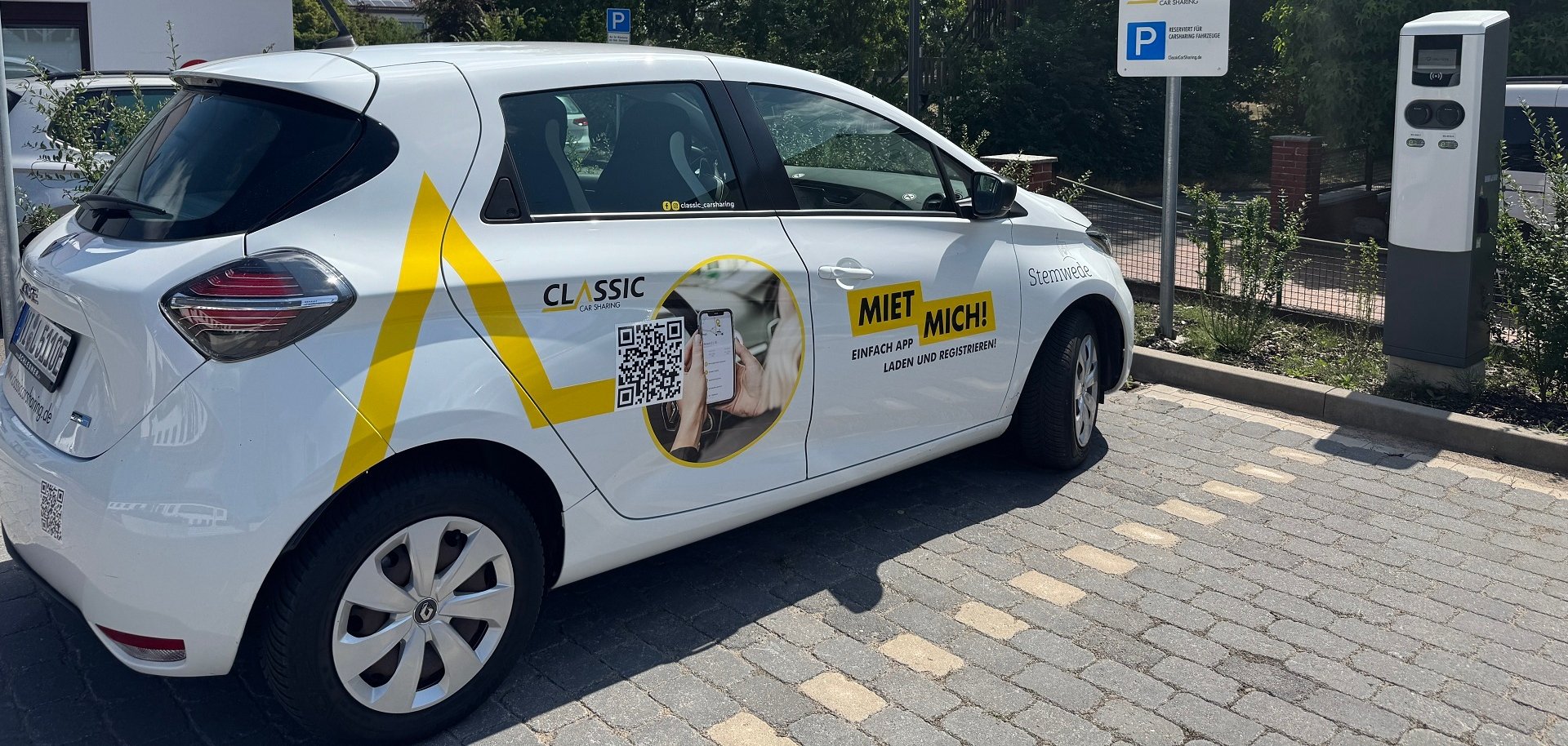 Ein weißes Carsharing-Fahrzeug steht auf einem ausgewiesenen Stellplatz neben einer Ladesäule. Am Fahrzeug befinden sich gut sichtbare Aufkleber mit dem Schriftzug „Classic Car Sharing“, einem QR-Code sowie dem Hinweis „Miet mich!“. Im Hintergrund sind ein Verkehrsschild für den Carsharing-Stellplatz, Bäume und ein Gebäude zu sehen. Die Aufnahme entstand bei sonnigem Wetter.