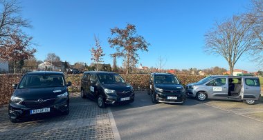 Seniorenfahrdienst Stemwede Vier Fahrzeuge des Seniorenfahrdienstes stehen auf einem Parkplatz. Drei schwarze und ein graues Fahrzeug sind nebeneinander geparkt, an den Fahrzeugen sind Logos des Fahrdienstes angebracht. Beim grauen Fahrzeug ist die seitliche Schiebetür geöffnet und gibt den Blick auf den Innenraum frei. Im Hintergrund sind Bäume, Wohnhäuser und eine Kirche unter blauem Himmel zu sehen.