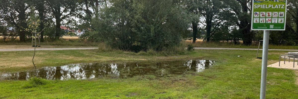 Vandalismus Kurpark Eine große Wasserlache bedeckt einen Teil der Rasenfläche im Kurpark nahe eines Spielplatzes. Rechts steht ein Schild mit der Aufschrift „Spielplatz“, im Hintergrund verlaufen Spazierwege vor einer Reihe hoher Bäume. Die Wasserfläche spiegelt die Bäume wider und zeigt, dass der Boden stark durchnässt ist. Links und im Hintergrund sind weitere Grünflächen sowie Spiel- und Bewegungsangebote des Kurparks zu erkennen.