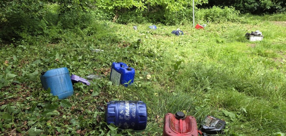 Altölbehälter im Stemweder Berg Mehrere Altölkanister und Ölbehälter liegen verstreut auf einer bewachsenen Fläche am Rand eines Naturschutzgebiets. Zwischen Gras, Brennnesseln und Sträuchern sind blaue, rote und schwarze Behälter zu erkennen. Im Hintergrund steht ein Schild, das auf das Naturschutzgebiet hinweist. Die Aufnahme zeigt die illegale Entsorgung von Altölbehältern in einem geschützten Naturbereich.
