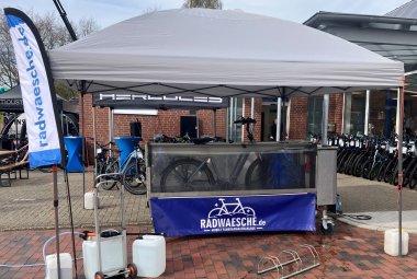Unter einem weißen Pavillon steht eine mobile Fahrradwaschanlage mit dem Aufdruck „Radwaesche.de“. Links daneben befindet sich eine Werbefahne. Im Hintergrund sind zahlreiche Fahrräder vor einem Fahrradgeschäft ausgestellt. Die Anlage steht auf einem gepflasterten Platz, daneben sind Wasserkanister und Zubehör für den Betrieb der Fahrradwäsche zu sehen. Das Foto zeigt einen Informations- oder Aktionstag rund ums Fahrrad.