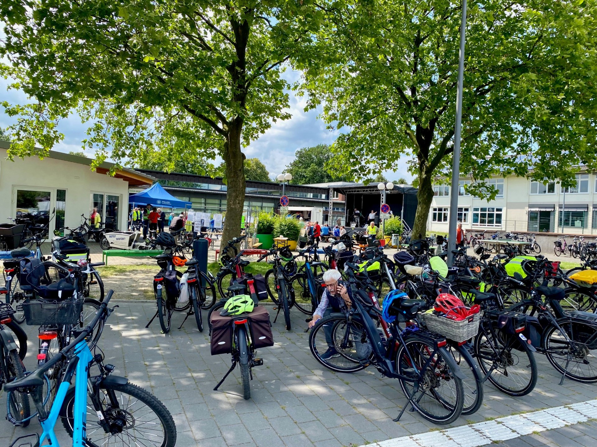 Auf einem Platz unter großen, schattenspendenden Bäumen stehen zahlreiche Fahrräder, viele davon mit Taschen, Körben oder Helmen ausgestattet. Im Hintergrund sind mehrere Menschen an Informationsständen und vor einer kleinen Bühne zu sehen. Eine Person überprüft ihr Fahrrad zwischen den abgestellten Rädern. Die Szene vermittelt den Eindruck einer gut besuchten Fahrradveranstaltung bei sonnigem Wetter.