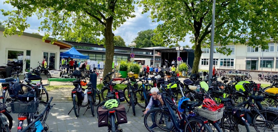 Auf einem Platz unter großen, schattenspendenden Bäumen stehen zahlreiche Fahrräder, viele davon mit Taschen, Körben oder Helmen ausgestattet. Im Hintergrund sind mehrere Menschen an Informationsständen und vor einer kleinen Bühne zu sehen. Eine Person überprüft ihr Fahrrad zwischen den abgestellten Rädern. Die Szene vermittelt den Eindruck einer gut besuchten Fahrradveranstaltung bei sonnigem Wetter.