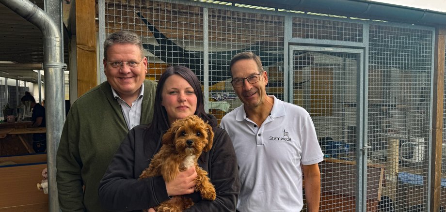 Tierheimbesuch Drei Erwachsene stehen vor einem Tiergehege mit Drahtgitter. Die Person in der Mitte hält einen kleinen braunen Hund mit lockigem Fell auf dem Arm. Links steht eine Person in grünem Pullover, rechts eine Person in weißem Poloshirt. Alle blicken in die Kamera. Im Hintergrund sind die Gehegeanlage, Holzelemente und ein überdachter Außenbereich zu sehen. Der Himmel ist bewölkt.