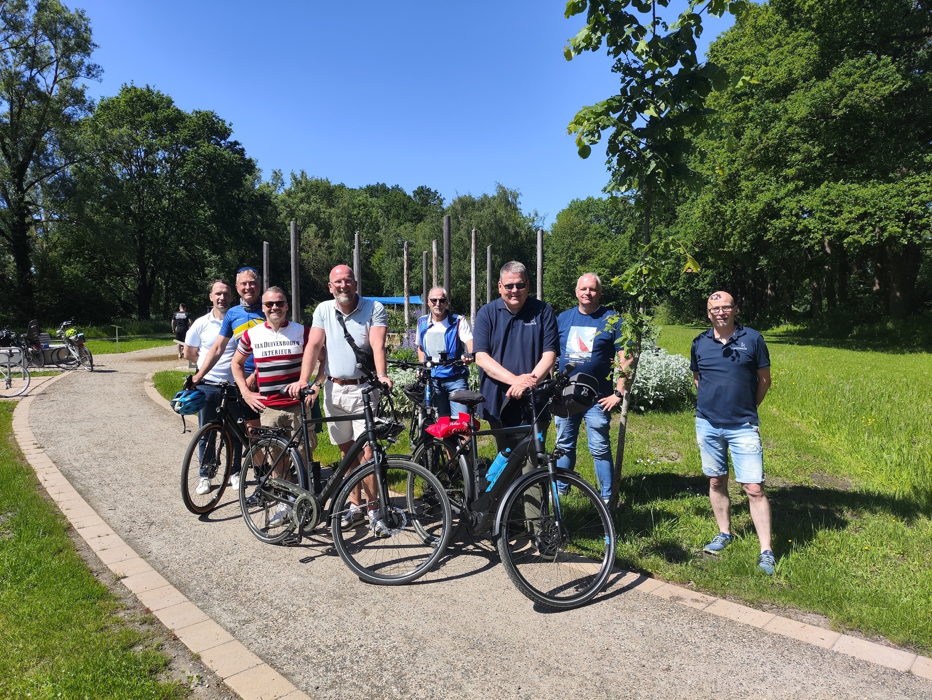 Acht Männer stehen mit mehreren Fahrrädern auf einem breiten, geschwungenen Weg in einer grünen Parkanlage. Die Gruppe blickt in die Kamera und posiert für ein gemeinsames Foto. Im Hintergrund sind hohe Bäume, eine gepflegte Rasenfläche und mehrere senkrechte Holzstelen zu sehen. Die Aufnahme entstand bei sonnigem Wetter unter klarem, blauem Himmel. Die Fahrräder stehen im Vordergrund, während die Teilnehmer der Radtour dahinter und daneben angeordnet sind. Die Umgebung wirkt ruhig, naturnah und einladend.