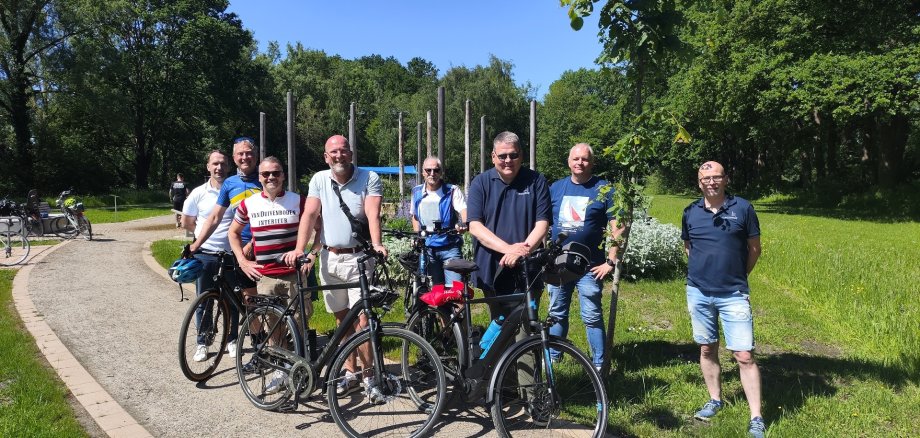 Acht Männer stehen mit mehreren Fahrrädern auf einem breiten, geschwungenen Weg in einer grünen Parkanlage. Die Gruppe blickt in die Kamera und posiert für ein gemeinsames Foto. Im Hintergrund sind hohe Bäume, eine gepflegte Rasenfläche und mehrere senkrechte Holzstelen zu sehen. Die Aufnahme entstand bei sonnigem Wetter unter klarem, blauem Himmel. Die Fahrräder stehen im Vordergrund, während die Teilnehmer der Radtour dahinter und daneben angeordnet sind. Die Umgebung wirkt ruhig, naturnah und einladend.
