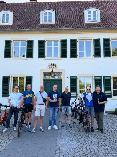 Sieben Männer stehen mit mehreren Fahrrädern vor einem historischen, hell verputzten Gebäude mit grünen Fensterläden und rotem Ziegeldach. Die Gruppe posiert für ein gemeinsames Foto auf dem gepflasterten Vorplatz vor dem Eingang. Über der Tür befindet sich ein Wappen. Einige Teilnehmer tragen Freizeitkleidung, einer ein Fahrradtrikot. Die Fahrräder stehen neben den Personen oder werden festgehalten. Das Foto entstand bei sonnigem Wetter, die Fenster spiegeln die umliegenden Bäume wider.