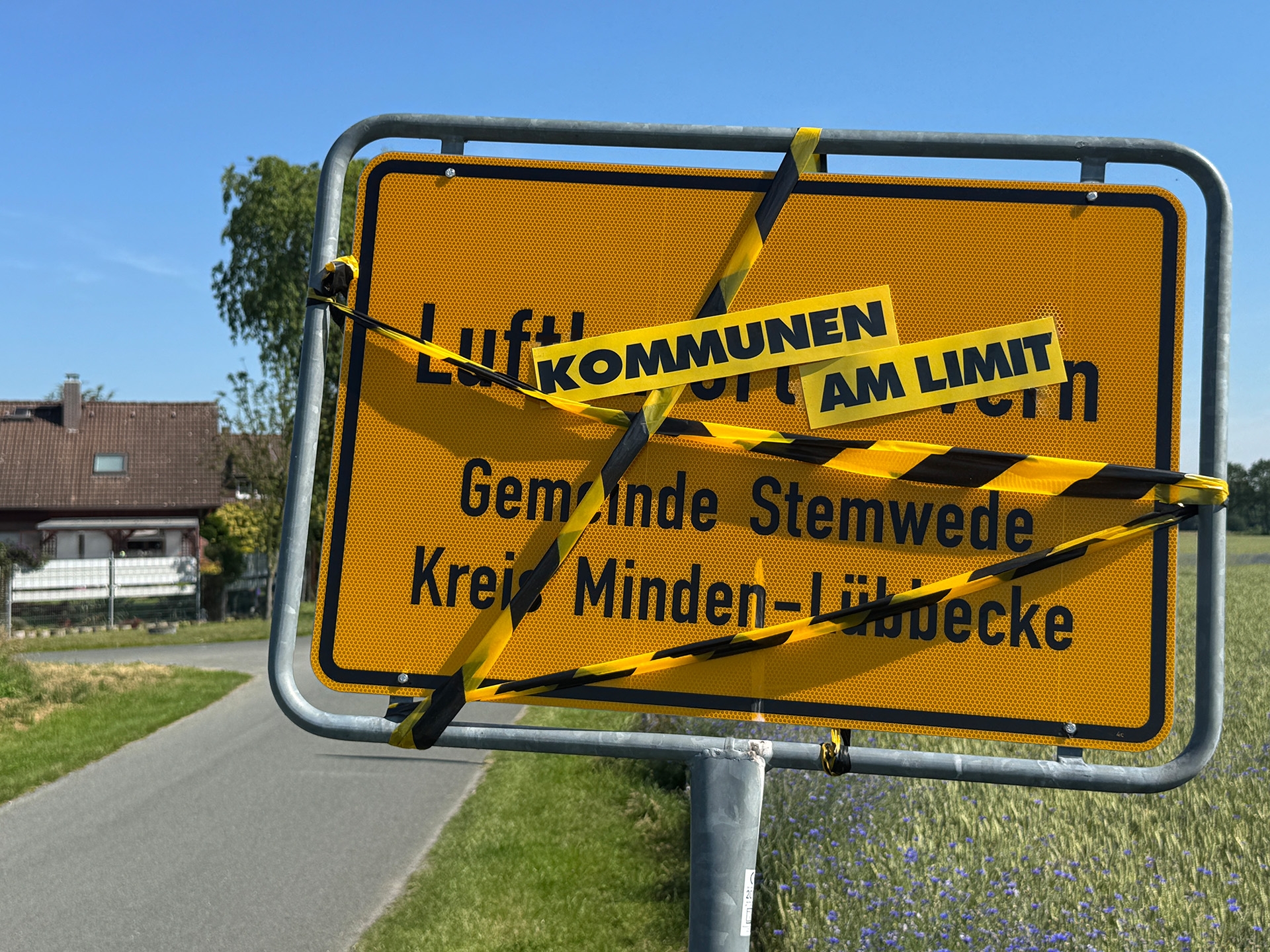 Ein gelbes Ortseingangsschild mit der Aufschrift „Luftkurort Wehdem, Gemeinde Stemwede, Kreis Minden-Lübbecke“ steht an einer Landstraße. Das Schild ist mit gelb-schwarz gestreiftem Absperrband umwickelt. Darüber sind die Worte „KOMMUNEN“ und „AM LIMIT“ auf gelben Schildern angebracht. Im Hintergrund sind Felder, ein Wohnhaus und ein klarer blauer Himmel zu sehen.