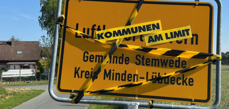 Ein gelbes Ortseingangsschild mit der Aufschrift „Luftkurort Wehdem, Gemeinde Stemwede, Kreis Minden-Lübbecke“ steht an einer Landstraße. Das Schild ist mit gelb-schwarz gestreiftem Absperrband umwickelt. Darüber sind die Worte „KOMMUNEN“ und „AM LIMIT“ auf gelben Schildern angebracht. Im Hintergrund sind Felder, ein Wohnhaus und ein klarer blauer Himmel zu sehen.