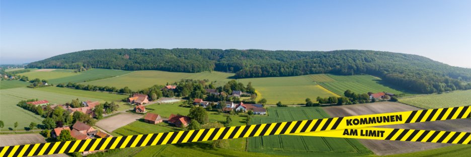 Luftaufnahme einer ländlichen Landschaft mit Feldern, Wiesen, einzelnen Höfen und kleinen Ortschaften vor einem bewaldeten Höhenzug unter klarem blauem Himmel. Im unteren Bildbereich verläuft ein gelb-schwarz gestreiftes Absperrband diagonal über das Bild. Auf dem Band steht in schwarzer Schrift „KOMMUNEN AM LIMIT“.