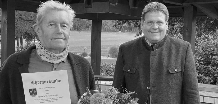 Ewald Kreienbrock Schwarz-Weiß-Fotografie von zwei Männern, die unter einem überdachten Pavillon stehen. Links hält ein älterer Mann eine Ehrenurkunde und einen Blumenstrauß in den Händen. Die Urkunde würdigt seine langjährige Tätigkeit als Mitglied des Gemeinderates und seine Ernennung zum Ehrenmitglied. Rechts steht ein weiterer Mann in einem dunklen Sakko und lächelt in die Kamera. Im Hintergrund sind eine Grünfläche, Bäume und Sträucher zu sehen. Die Aufnahme vermittelt den Eindruck einer feierlichen Ehrung und Anerkennung für langjähriges kommunalpolitisches Engagement.