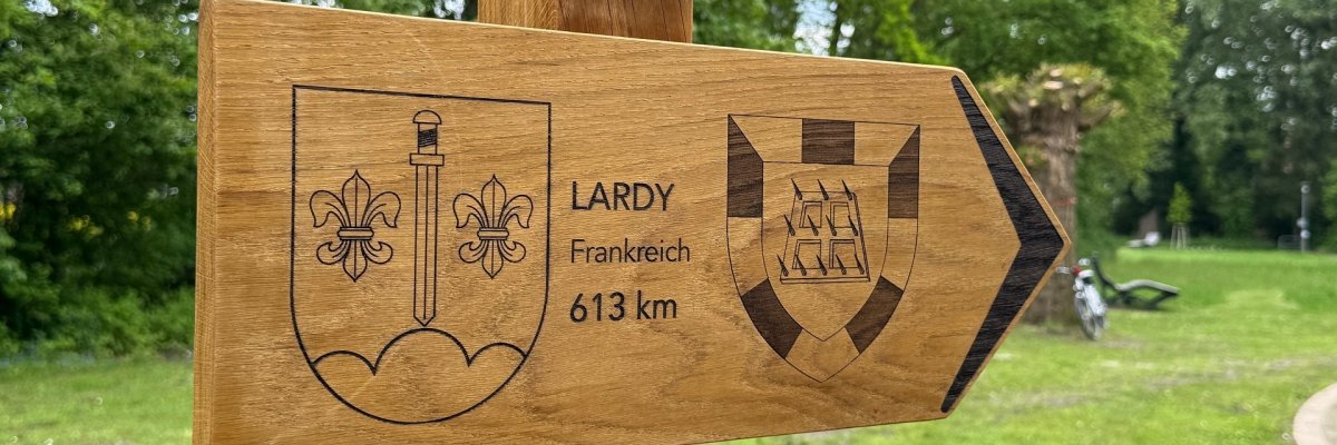 Partnerschaftsschild Ein hölzernes Wegweiserschild in Form eines Pfeils ist an einem Holzpfosten befestigt und zeigt nach rechts. Auf dem Schild sind zwei Wappen eingraviert. Dazwischen steht die Aufschrift „LARDY“, darunter „Frankreich“ und „613 km“. Das Schild befindet sich in einer grünen Parklandschaft mit Bäumen und Rasen. Im unscharfen Hintergrund sind rechts ein Fahrrad und eine Bank zu erkennen.
