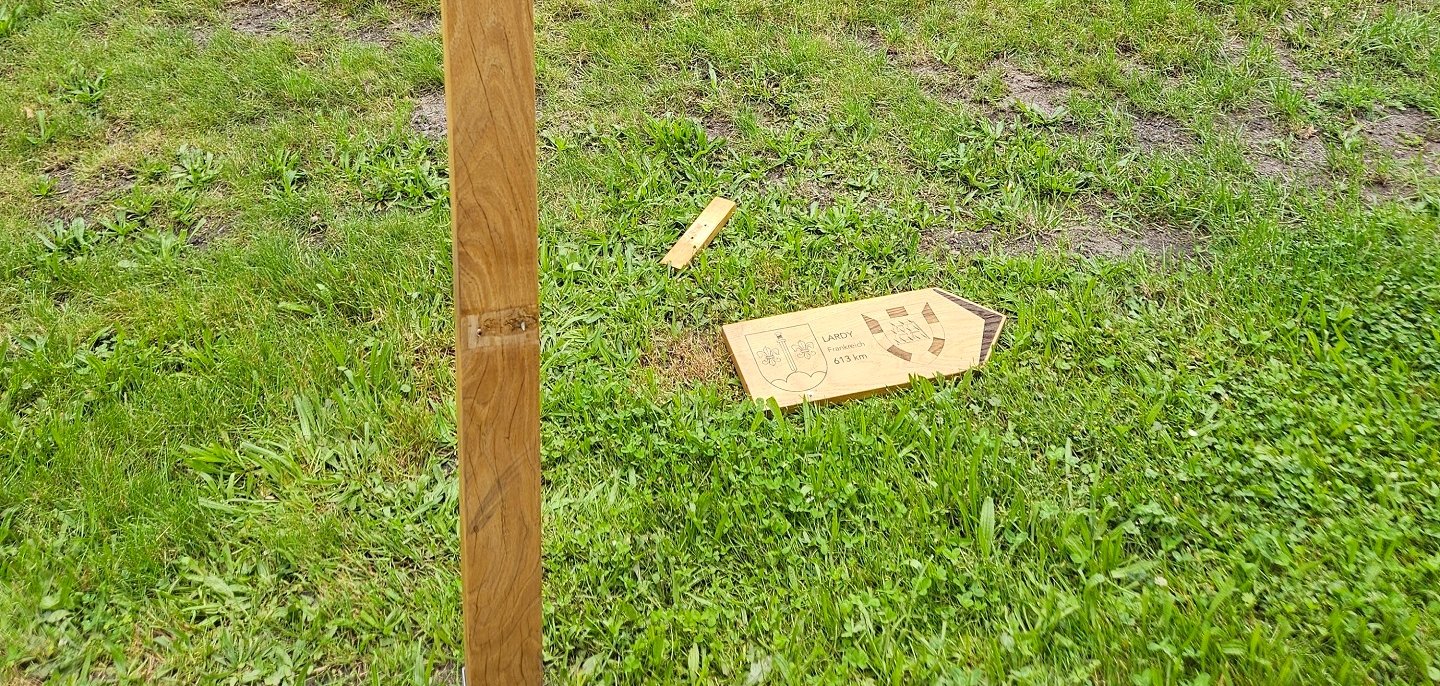 Ein hölzerner Pfosten steht auf einer Rasenfläche in einem Park. Das dazugehörige Wegweiserschild liegt abgebrochen im Gras neben dem Pfosten. Das Schild trägt die Aufschrift „LARDY, Frankreich, 613 km“ und zeigt zwei Wappen. Im Hintergrund sind Bäume, Sträucher und eine offene Grünfläche zu sehen. Die Szene dokumentiert eine Beschädigung oder einen Vandalismusschaden an der Beschilderung.