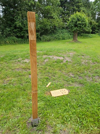 Vandalismus Ein hölzerner Pfosten steht auf einer Rasenfläche in einem Park. Das dazugehörige Wegweiserschild liegt abgebrochen im Gras neben dem Pfosten. Das Schild trägt die Aufschrift „LARDY, Frankreich, 613 km“ und zeigt zwei Wappen. Im Hintergrund sind Bäume, Sträucher und eine offene Grünfläche zu sehen. Die Szene dokumentiert eine Beschädigung oder einen Vandalismusschaden an der Beschilderung.