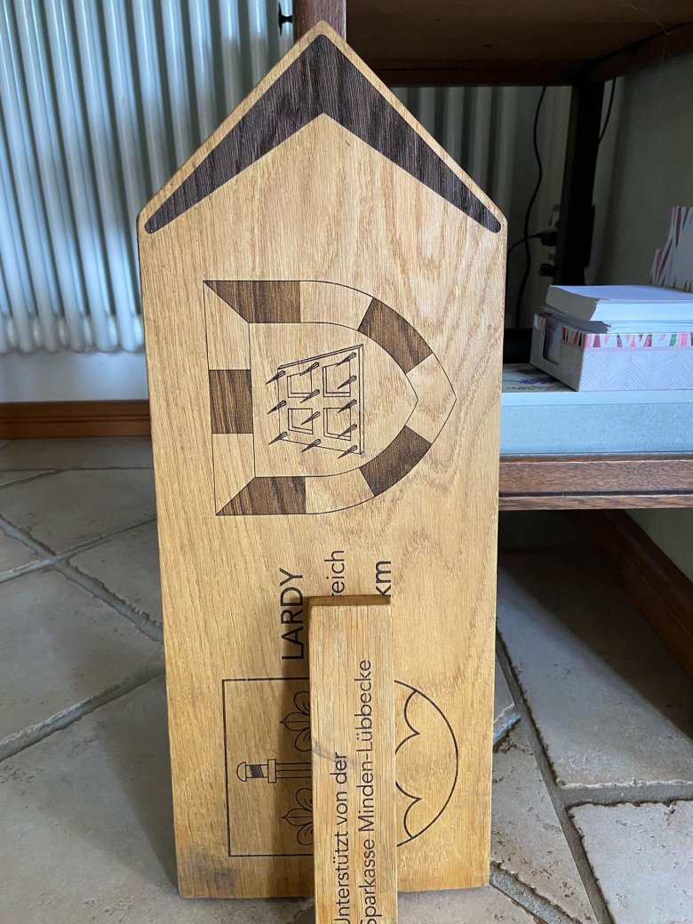 Vandalismus Ein beschädigtes hölzernes Wegweiserschild lehnt in einem Innenraum an einem Möbelstück. Das Schild zeigt die Aufschrift „LARDY“, Teile der Angaben „Frankreich“ und „613 km“ sowie zwei eingravierte Wappen. Ein abgebrochenes Holzstück liegt vor dem Schild und verdeckt einen Teil der Beschriftung. Auf dem Holzstück steht „unterstützt von der Sparkasse Minden-Lübbecke“. Die Aufnahme dokumentiert den Schaden am Schild nach einer mutmaßlichen mutwilligen Beschädigung.