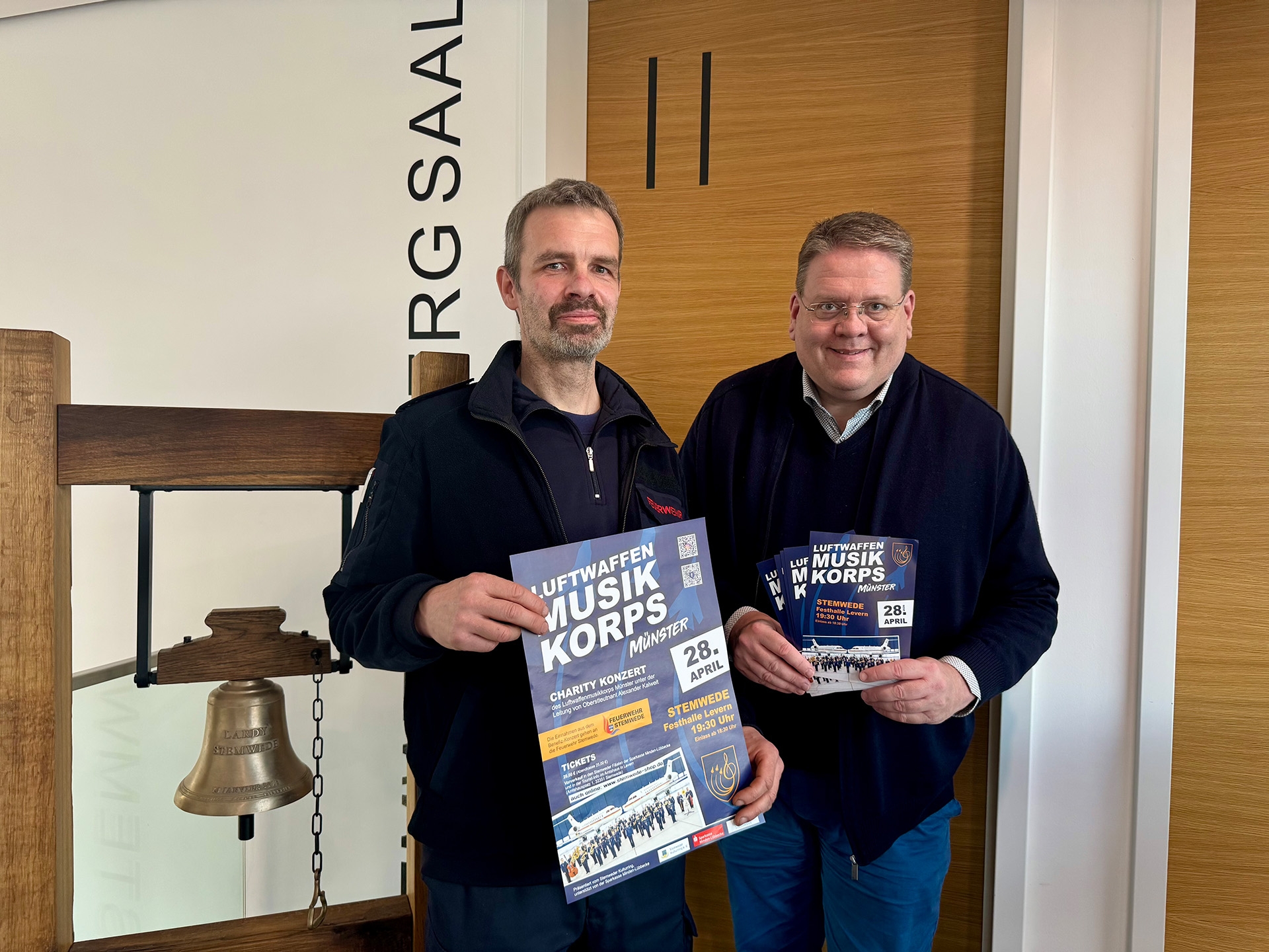 Zwei Männer stehen in einem Innenraum neben einer großen, freihängenden Glocke aus Metall in einem Holzgestell. Beide halten Plakate bzw. Flyer mit der Aufschrift „Luftwaffen Musikkorps Münster“. Auf dem Plakat steht unter anderem „Charity Konzert“ sowie das Datum „28. April“; weitere Angaben zu Ort und Uhrzeit sind abgedruckt. Im Hintergrund sind eine Holztür und eine Wand mit teilweise sichtbarer Beschriftung zu sehen.